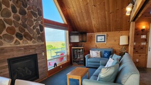 A cozy living room with a stone wall, a fireplace, a large window with a view, a blue sofa, and a wooden ceiling.