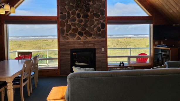 A cozy living room with a stone fireplace, large windows offering ocean views, a dining area with chairs, and a television.