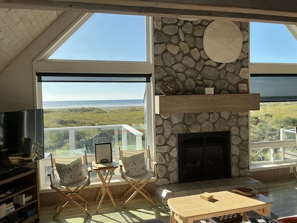 A spacious living area with a stone fireplace, large windows offering a view of the beach, and a television on the left.