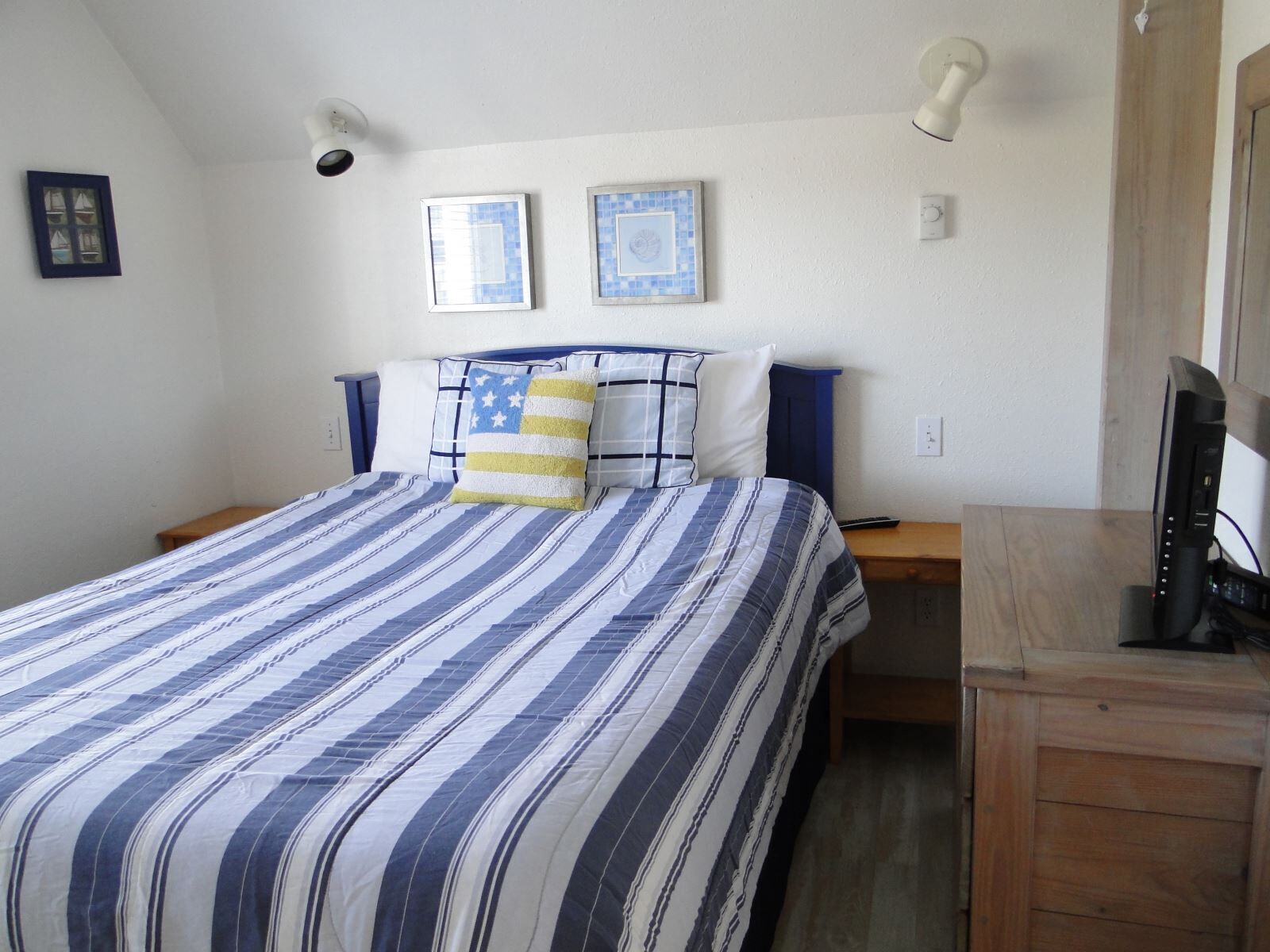 A bedroom with a bed, two wall-mounted lights, a small wooden nightstand, a television on a wooden stand, and two framed pictures on the wall.