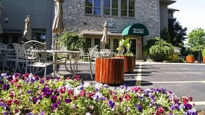 A stone building with a bar and grill sign, outdoor seating with umbrellas, and a flower bed in front.