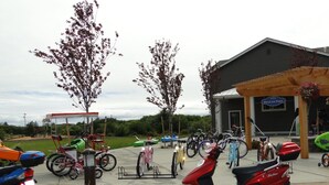 A parking area with various bicycles and a red scooter. There are trees and a building labeled "Pelican Post" in the background. - Outlook 309/Oyhut Bay (Ocean Shores)