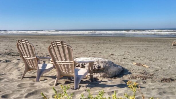 Two Adirondack chairs and a small table on a sandy beach with the ocean in the background.