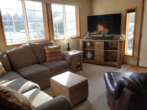 A living room with a sofa, ottoman, and leather chair. There is a TV on a wooden stand, a lamp, and a clock. - Clam Xing- Just blocks to the beach (Hoquiam)