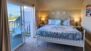 Quarto com cama, duas mesas de cabeceira com abajures, vista para o exterior por uma porta de correr e uma pequena varanda.