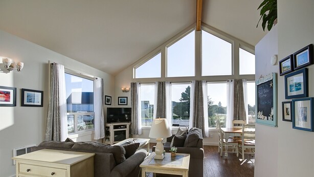 A spacious living room with a large window, a television, a sofa, a table, and a chair. - Sand Dollar- Adorable 2 bedroom  with a Hot tub, fenced yard, BBQ and pets ok . (Ocean Shores)