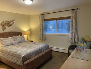 A bedroom with a bed, a nightstand, a window with curtains, a decorative wall piece, and a glass table with a decorative item. - Joe Creek Inn- Unique Home (Pacific Beach)