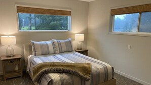 A neatly arranged bedroom with a bed, two bedside tables, lamps, and a window with a view of trees. - Joe Creek Inn- Unique Home (Pacific Beach)