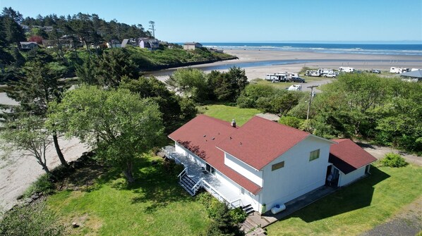 A single-story house with a red roof, a front porch, and a small deck. It is surrounded by greenery and located near a beach with RVs parked nearby. - Joe Creek Inn- Unique Home (Pacific Beach)