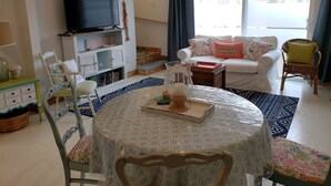 Een woonkamer met een televisie, een bank, een stoel, een tafel met tafelkleed en een raam met gordijnen.