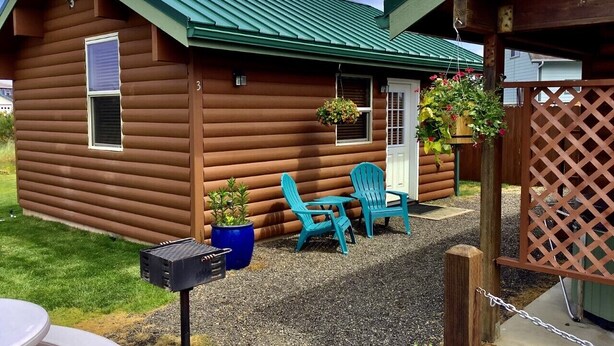 A log cabin with a green roof, a front door, and a small porch area with two turquoise chairs and a table. There are potted plants and a barbecue grill outside. - Moss Landing # 3 Studio (Ocean Shores)