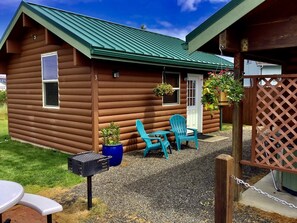 A log cabin with a green roof, a front door, and a small porch area with two turquoise chairs and a table. There are potted plants and a barbecue grill outside.