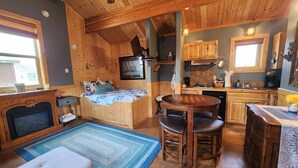 A cozy cabin interior with a wooden ceiling, a fireplace, a bed with a blue and white patterned bedspread, a dining table with four chairs, and a kitchen area with wooden cabinets and a stove. - Moss Landing # 3 Studio (Ocean Shores)