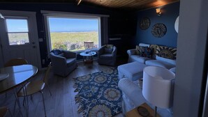 A cozy living room with a wooden ceiling, a large window offering a view of the outdoors, a patterned rug, and a sofa with cushions. - Moclips Sand Dollar Beach House - Beautiful Beach Front Home (Moclips)