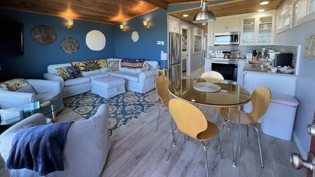 A modern living room with a glass dining table, a sofa set, and a kitchen area with white cabinets and stainless steel appliances. - Moclips Sand Dollar Beach House - Beautiful Beach Front Home (Moclips)