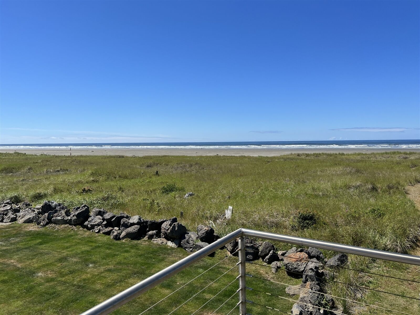 A grassy field with a metal railing in the foreground, leading to a beach with waves in the background.