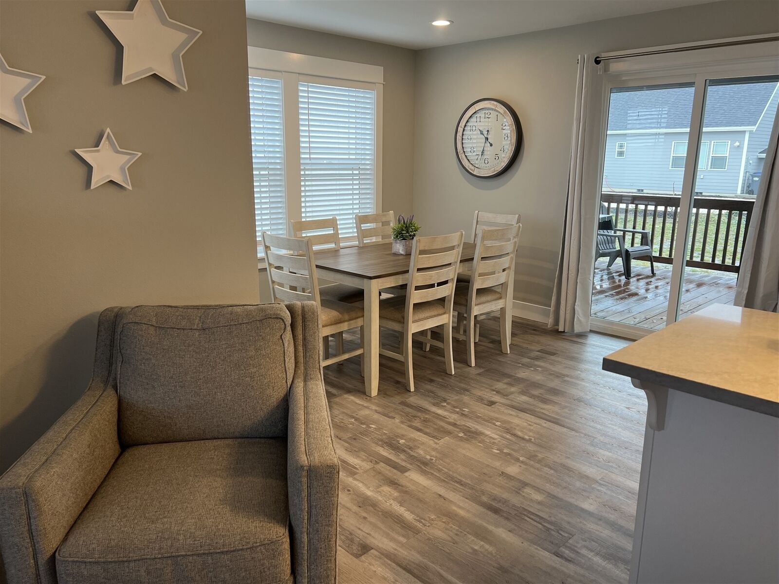 A dining area with a wooden table and chairs, a clock on the wall, and a view of the outside deck through a glass door.