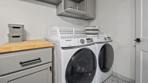 Un cuarto de lavado con lavadora de carga frontal, secadora, mostrador de madera, gabinetes grises y piso de baldosas blancas.