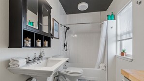 Un baño con lavamanos, inodoro y ducha. Hay estantes con artículos de aseo y una ventana con persianas.