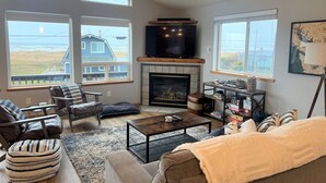 Ein geräumiges Wohnzimmer mit Kamin, großen Fenstern, einem Fernseher und Blick aufs Meer.