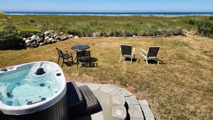 A hot tub with a stone deck, two chairs, and a table in a grassy area with a view of the ocean. - Beachcombers Cabin - Ocean Front with Hot Tub (Moclips)