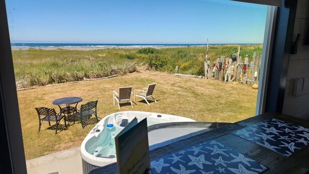 A view from a room with a hot tub and a patio area with chairs and a table, overlooking a beach and grassy area. - Beachcombers Cabin - Ocean Front with Hot Tub (Moclips)