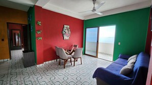 A living room with a red and green color scheme, a glass table with chairs, a blue sofa, and a sliding glass door leading to a balcony. - Velyn (Yelapa)