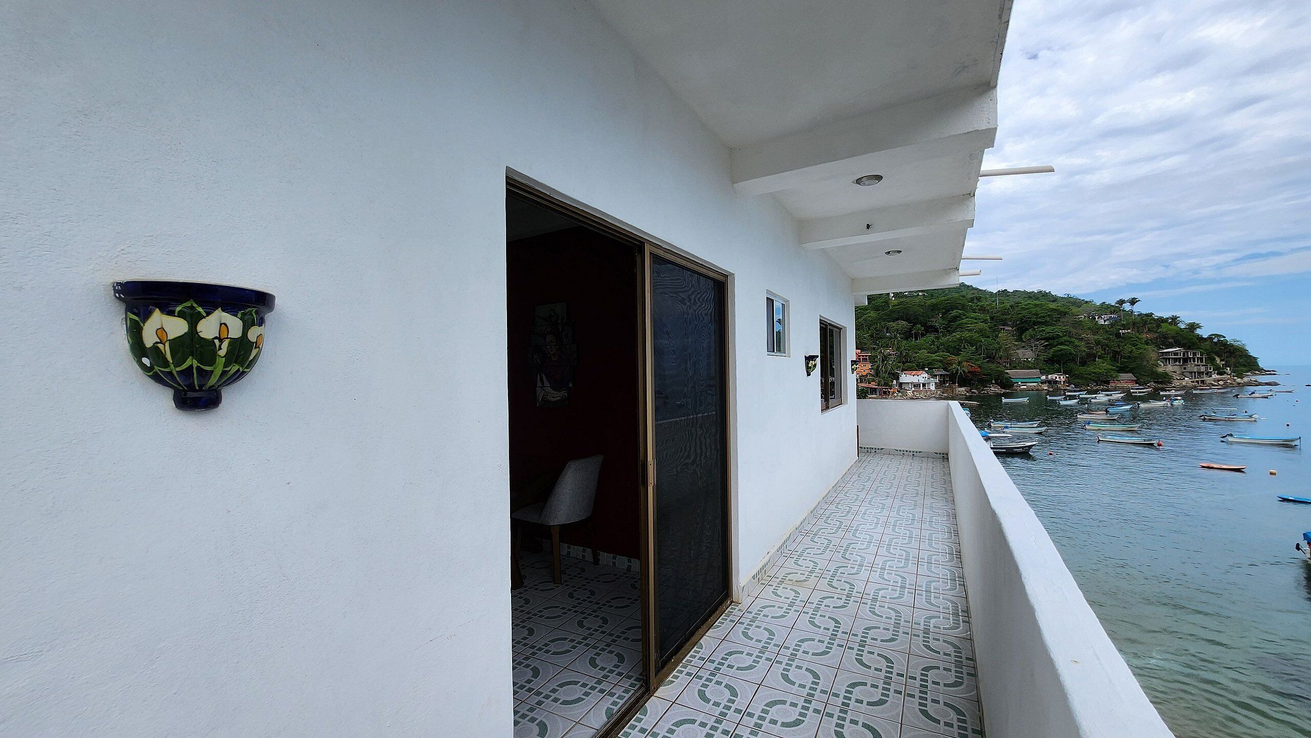 Un balcón con baldosas estampadas, una pared blanca con un adorno floral decorativo, y vistas a barcos en el agua.