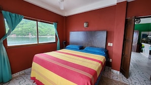 A bedroom with a bed, curtains, a view of a lake, and a door leading to a kitchen. - Velyn (Yelapa)