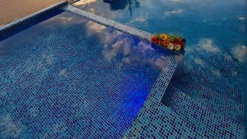 Piscina con borde de azulejos y arreglo floral, con vista a una playa con palmeras y montañas al fondo.