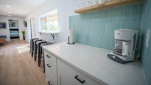 A modern kitchen with a coffee machine, bar stools, and a window overlooking a garden. - Contained in Gruene! Hot Tub! (New Braunfels)