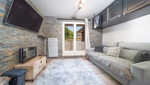 A modern living room with a stone wall, a flat-screen TV, a sofa with patterned cushions, and a fluffy rug. - Studio 2/4 ppl near the ski runs (Allos)