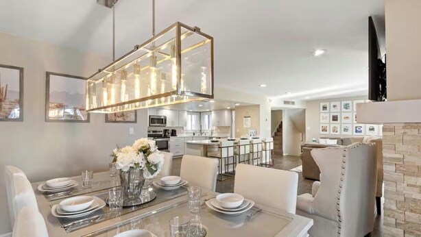 A modern dining area with a glass table set for six, a chandelier, and a kitchen in the background. -  🌴2 King Suites, Huge HEATED POOL& SPA, 8 MIN TO OLD TOWN,2 LIVING ROOMS, FIREPIT (Scottsdale)