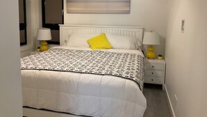 A neatly made bed with a white headboard, a single yellow pillow, and a floral-patterned bedspread. There are two bedside lamps with yellow bases and white shades. A painting of a landscape hangs above the bed.