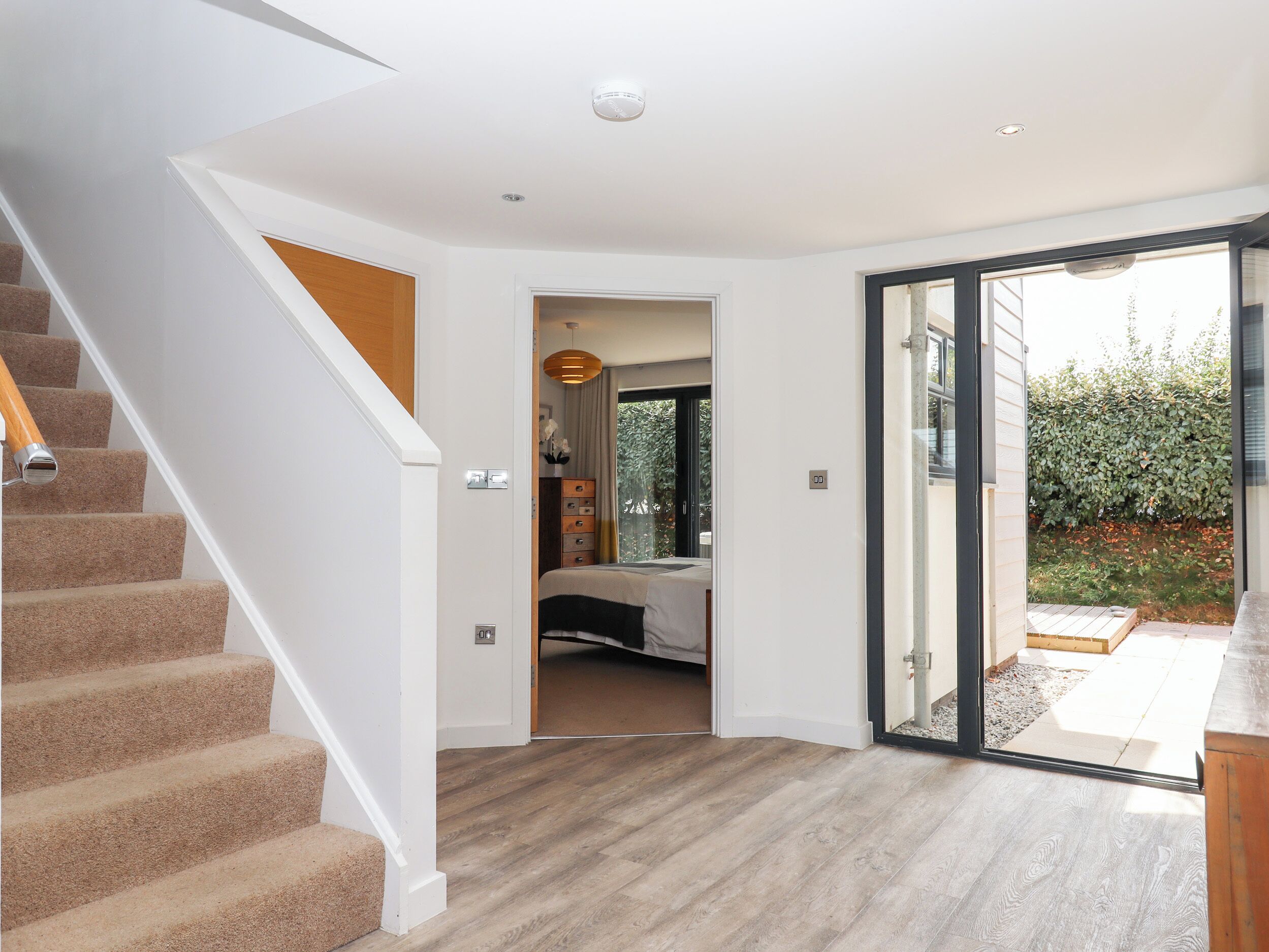 A modern interior with a staircase, a bedroom with a bed and dresser, and a sliding glass door leading to a garden.