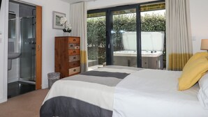 Ein Schlafzimmer mit einem großen Bett, einer hölzernen Kommode, einer Duschkabine aus Glas und Blick auf einen Garten durch eine Glastür.