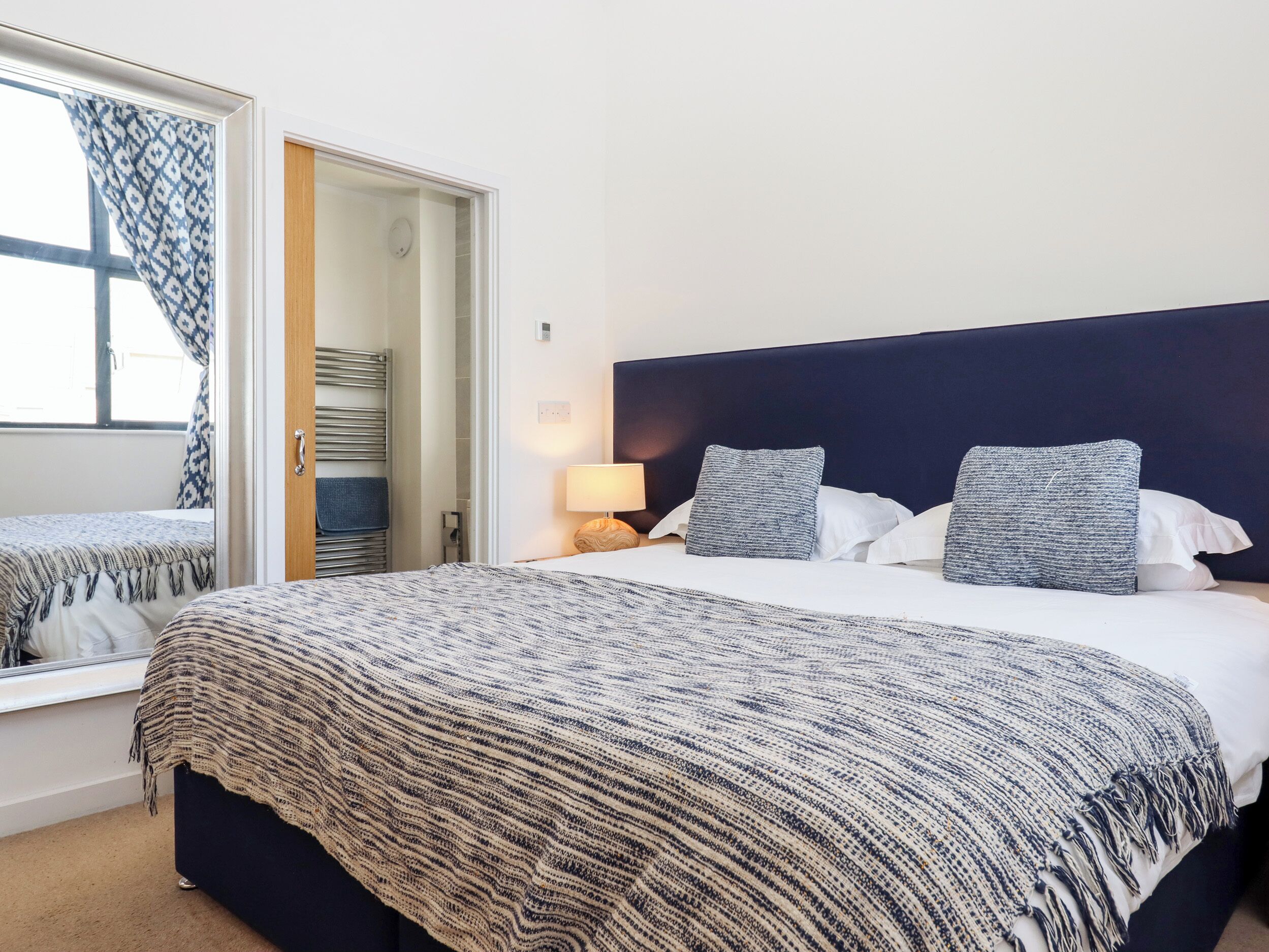 Una habitación de hotel con una cama grande, una mesita de noche con lámpara, cabecero y ventana con cortinas estampadas.