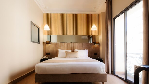 A modern hotel room with a large bed, wooden panel wall, two bedside lamps, and a large window with a balcony view. - Ennasma (Casablanca)
