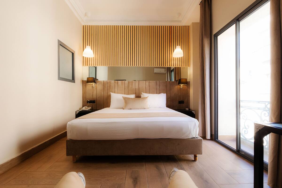 A modern hotel room with a large bed, wooden panel wall, two bedside lamps, and a large window with a balcony view.