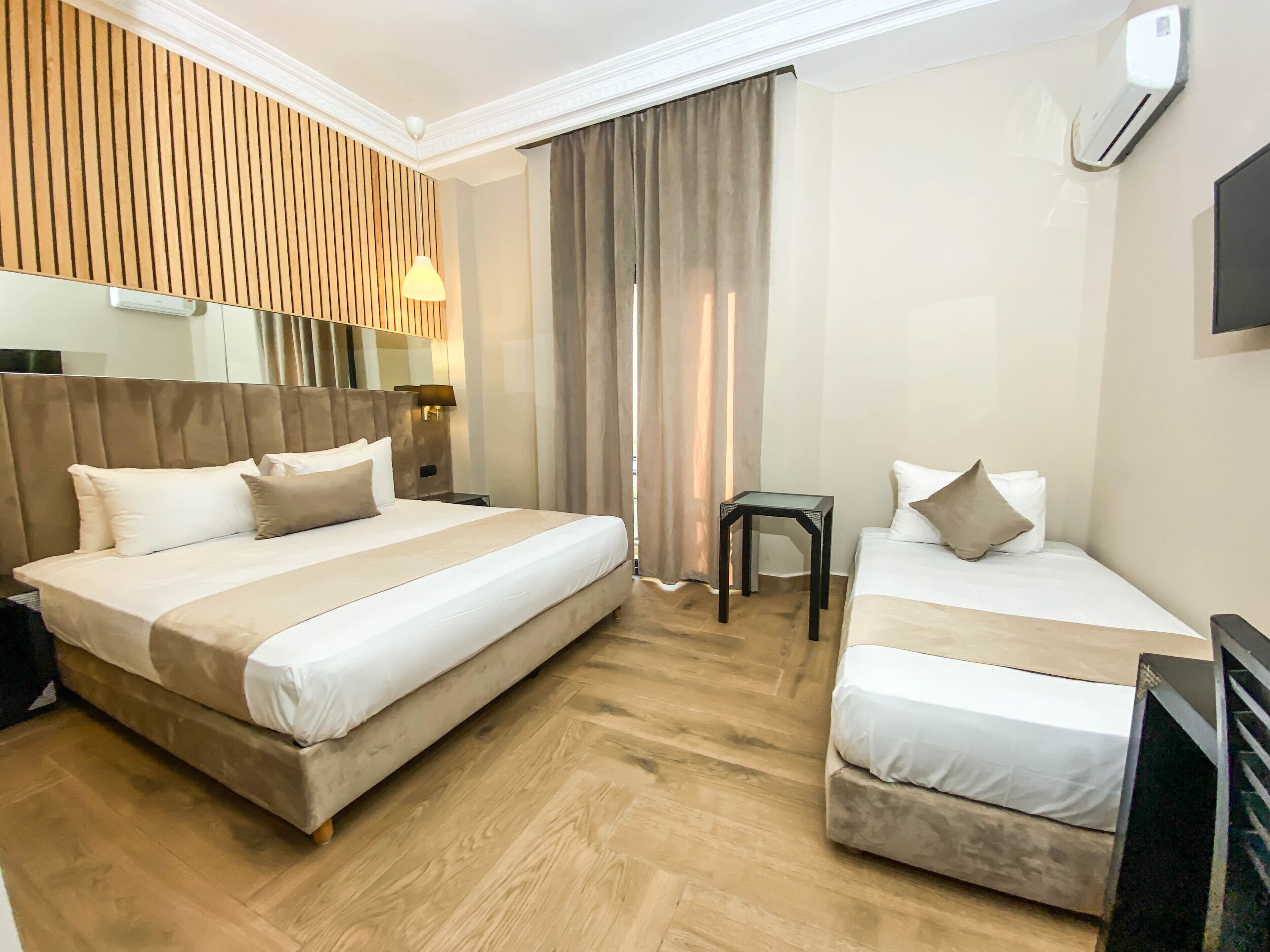 A hotel room with two beds, a wooden headboard, a small table, a TV, and a window with curtains.