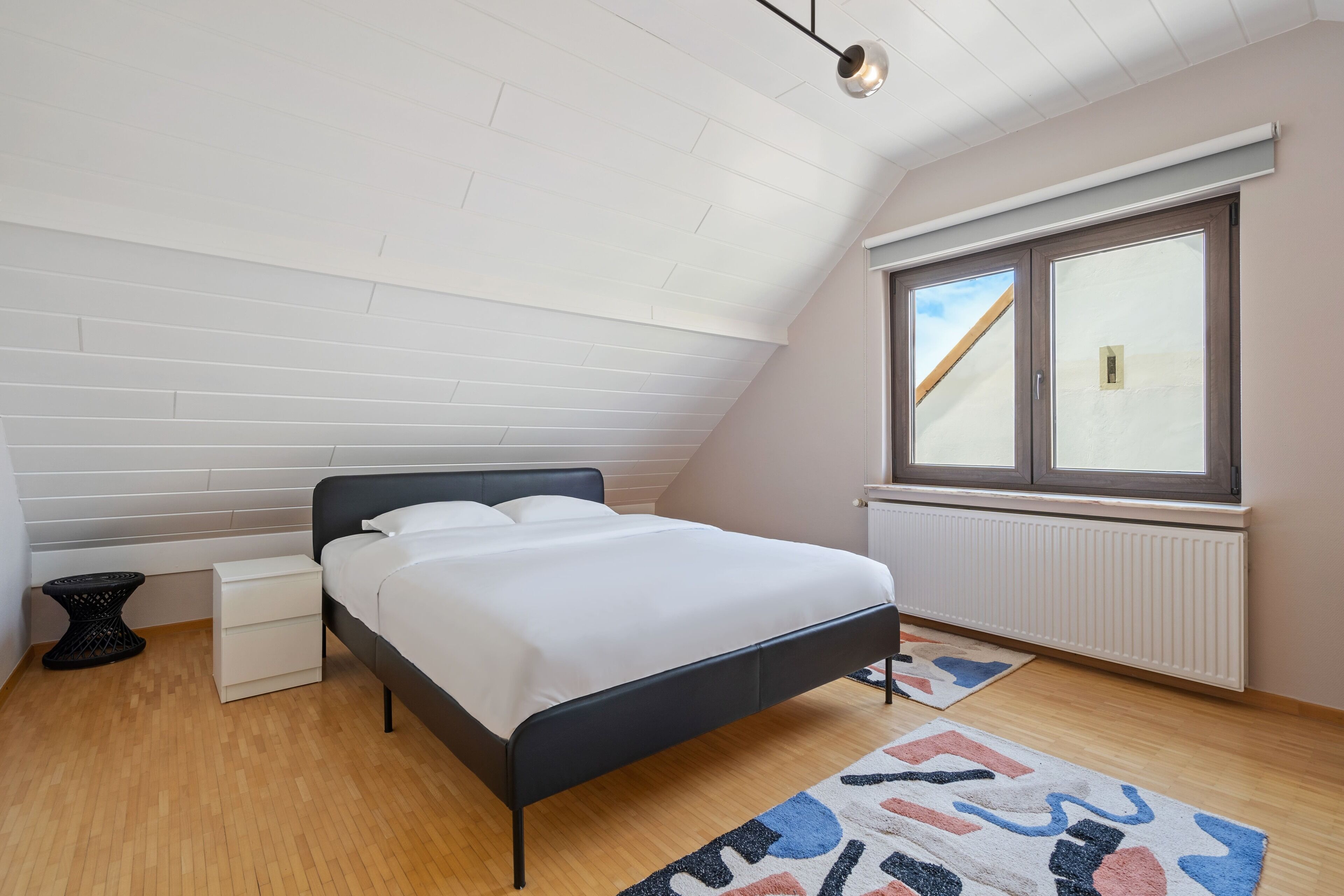 A bedroom with a bed, a window, a radiator, and a ceiling with wooden panels.