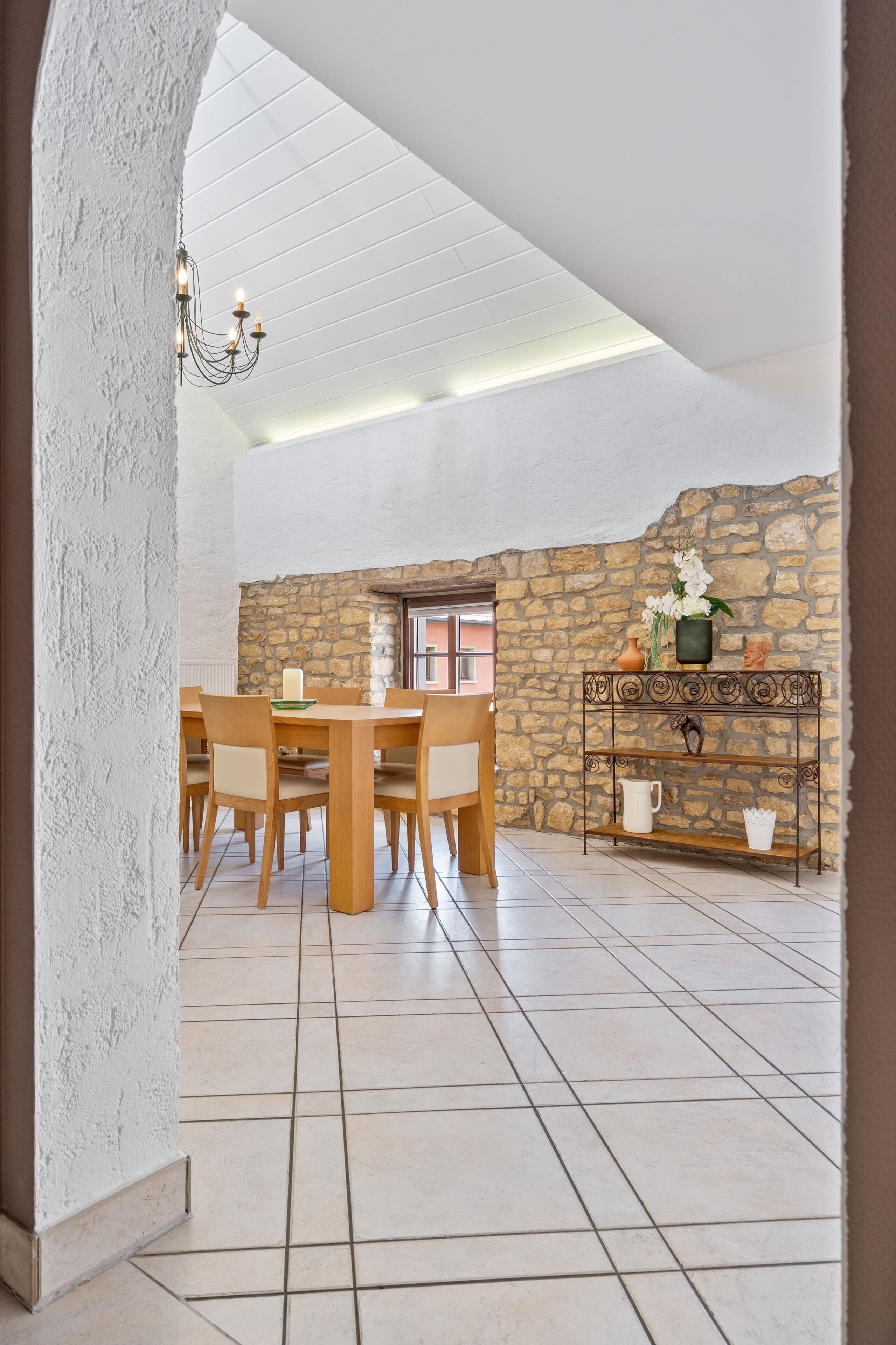 A dining area with a stone wall, a wooden table with chairs, a metal console table with decorative items, and a chandelier.