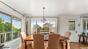 Comedor con mesa de madera, cuatro sillas, una araña y ventanas amplias con vista a follaje.
