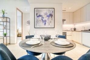 A modern dining area with a round table set for two, surrounded by blue chairs. A large abstract painting hangs above the table, and a kitchen is visible in the background. - Modern 2BR w/ Sea View Balcony at Address JBR (Dubai)