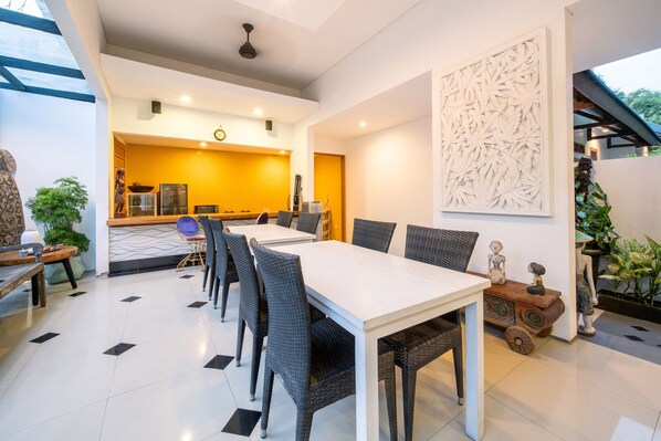 Un espace repas moderne avec une table blanche et des chaises en osier, une cuisine en arrière-plan et un élément décoratif mural.