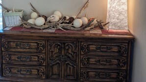 Uma cômoda de madeira com um quadro floral emoldurado acima, um cesto, ovos e galhos decorativos.