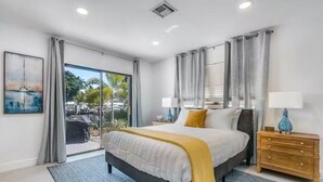 A modern bedroom with a large bed, a nightstand, a lamp, and a view of the outdoors through a sliding door.