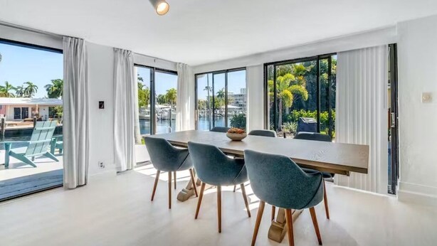 A modern dining area with a large table and four chairs, large windows with a view of palm trees and buildings, and a sliding glass door leading to an outdoor area with chairs.