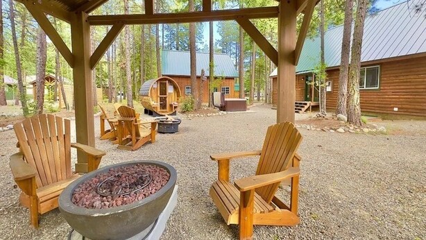 A covered outdoor seating area with Adirondack chairs and a fire pit, surrounded by trees and a cabin in the background.