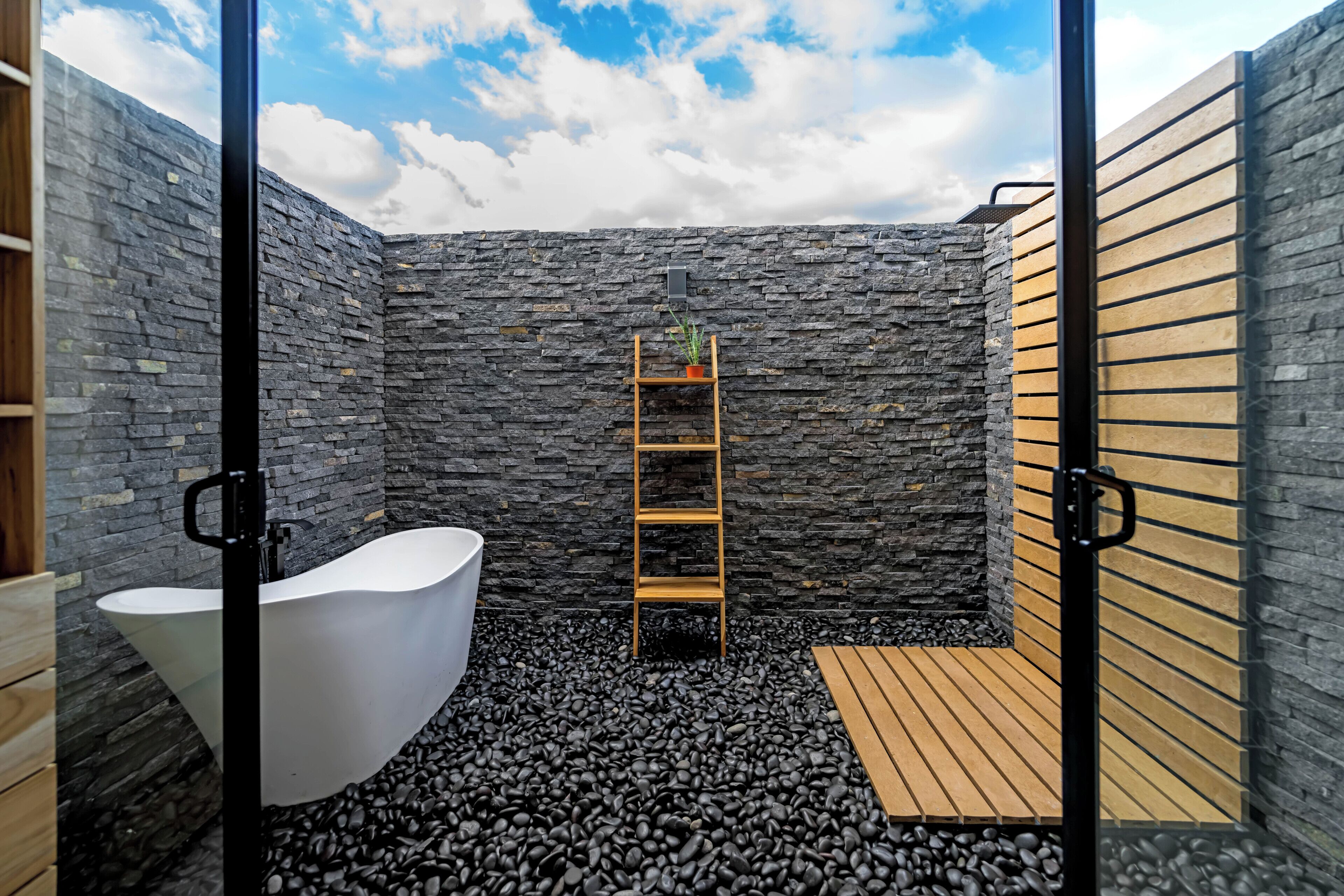 A modern bathroom with a stone wall, a white bathtub, a wooden ladder shelf, and a wooden floor.